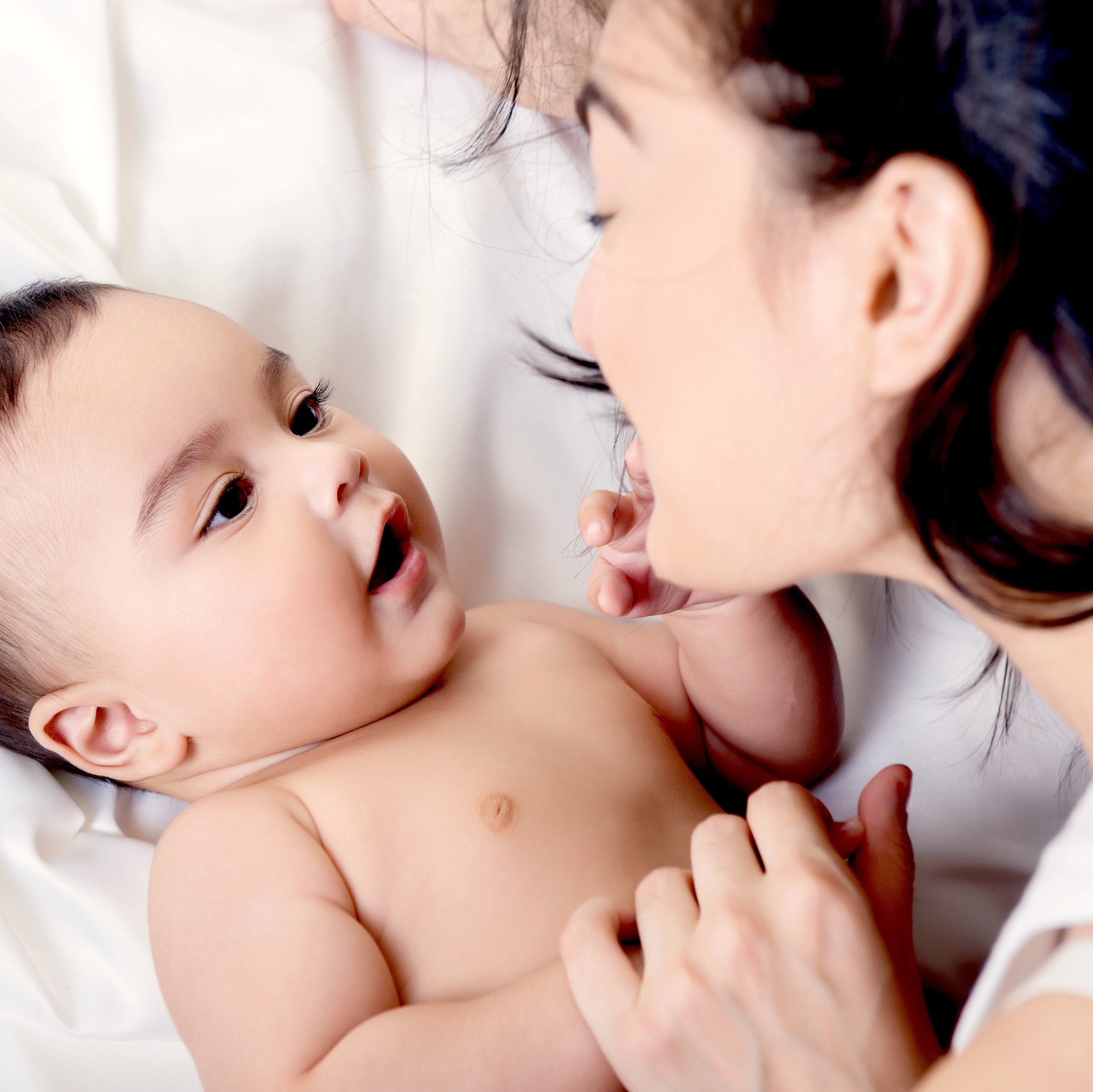 Young mother playing with her baby on white silk bed sheet. Cute mixed race bab boy has fun with mum, high key.